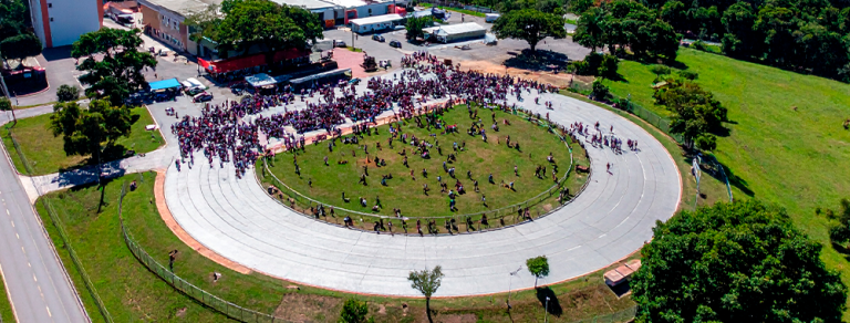 Vista aérea de evento escolar ao ar livre com grande número de pessoas reunidas em pista circular dentro de uma instituição.