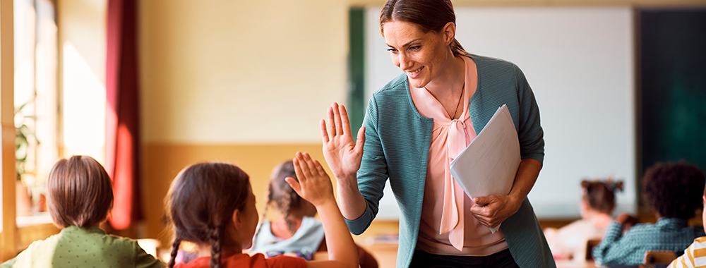 Professora sorridente cumprimenta aluna com um "high five" em sala de aula, simbolizando uma relação de respeito e valorização no ambiente escolar.