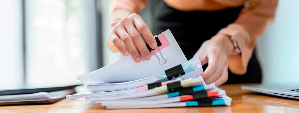 Pessoa organizando uma pilha de documentos e relatórios com clipes e marcadores, ilustrando a gestão de inadimplência escolar.