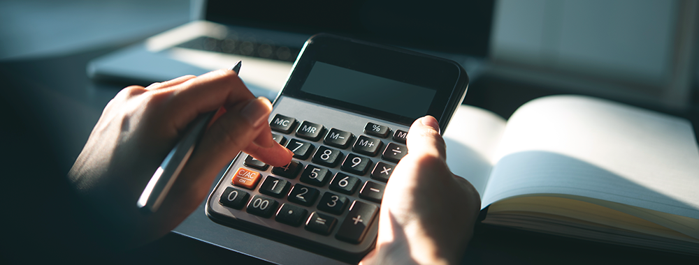 Mãos usando uma calculadora ao lado de um caderno e um laptop, ilustrando o cálculo de adicional noturno, insalubridade e periculosidade na folha de pagamento escolar.