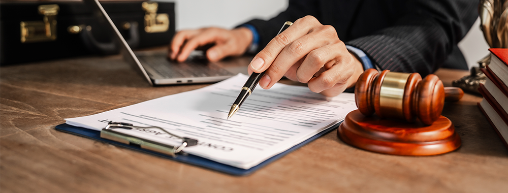 Mãos assinando um documento com caneta ao lado de um martelo de juiz, ilustrando compliance e prevenção de litígios trabalhistas.